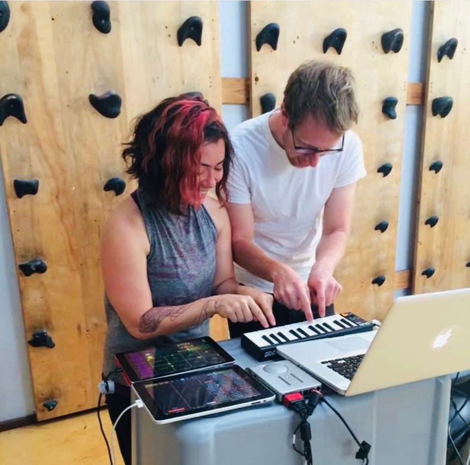 Two people making music together on a tiny keyboard and iPads in front of a climbing wall